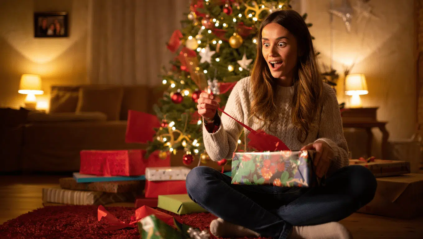 cadeau noel femme ouvrant son cadeau de Noël au pied du sapin dans son salon chaleureux et décoré pour les fêtes
